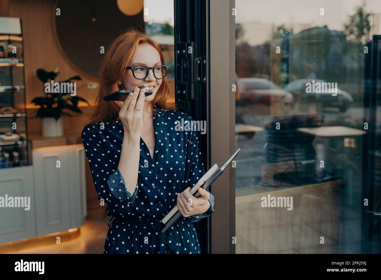 Lächelnde, nachdenkliche, rothaarige junge Frau telefoniert per Smartphone sieht weg Stände in der Tür des Cafés verwendet moderne Technologien für die Kommunikation Stockfoto