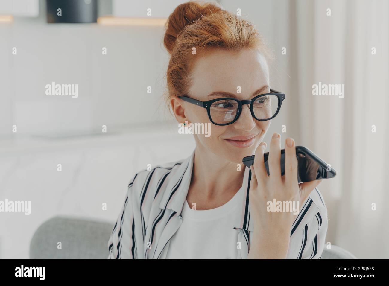 Horizontale Aufnahme von Ingwer Frau verwendet Sprachassistent auf Smartphone lässt Sprachanrufe aussehen hat Freude Ausdruck trägt transparente Brille Freizeit Stockfoto