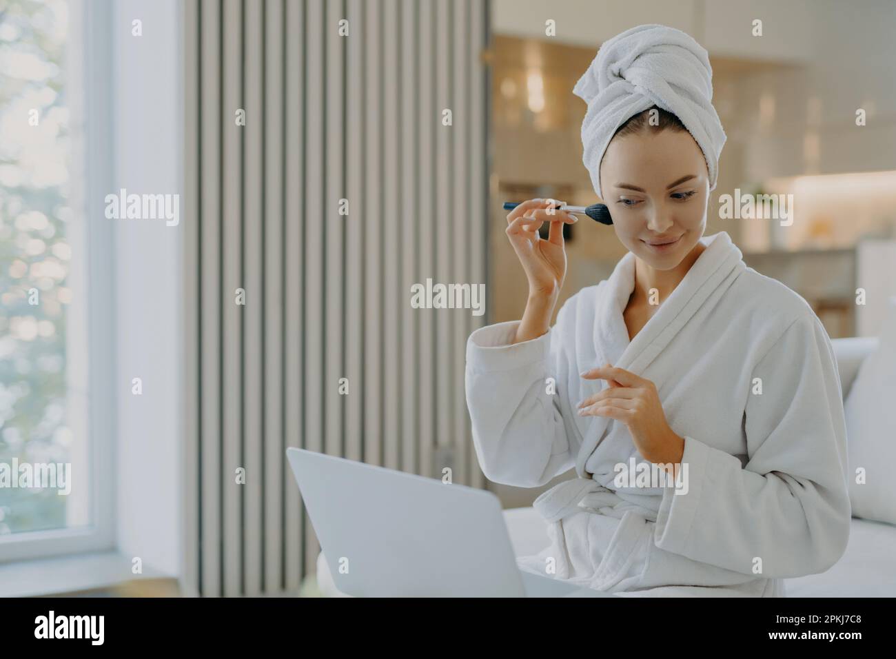 Foto einer jungen europäischen Frau trägt Gesichtspulver mit Kosmetikbürste auf, konzentriert in Laptop-Computer macht Videoanrufe und hat eigene Beauty-Blog-Posen in CO Stockfoto