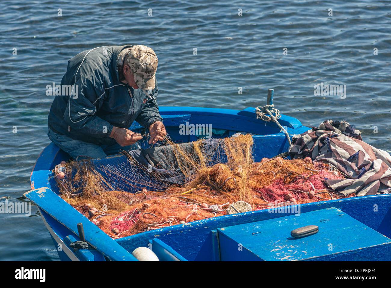 Ein Fischer mit einem Hut, der mit einer großen Nadel ein Fischernetz auf einem alten blauen hölzernen Fischerboot am Hafenkai in Bari, Apulien, Italien, repariert Stockfoto