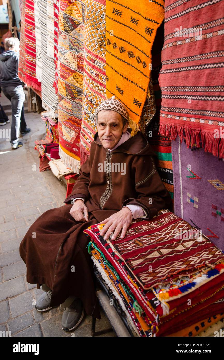 Ein Mann in traditioneller marokkanischer Djellaba und kufi-Hut sitzt neben seinen Teppichen zum Verkauf in Fez Medina Marokko Stockfoto