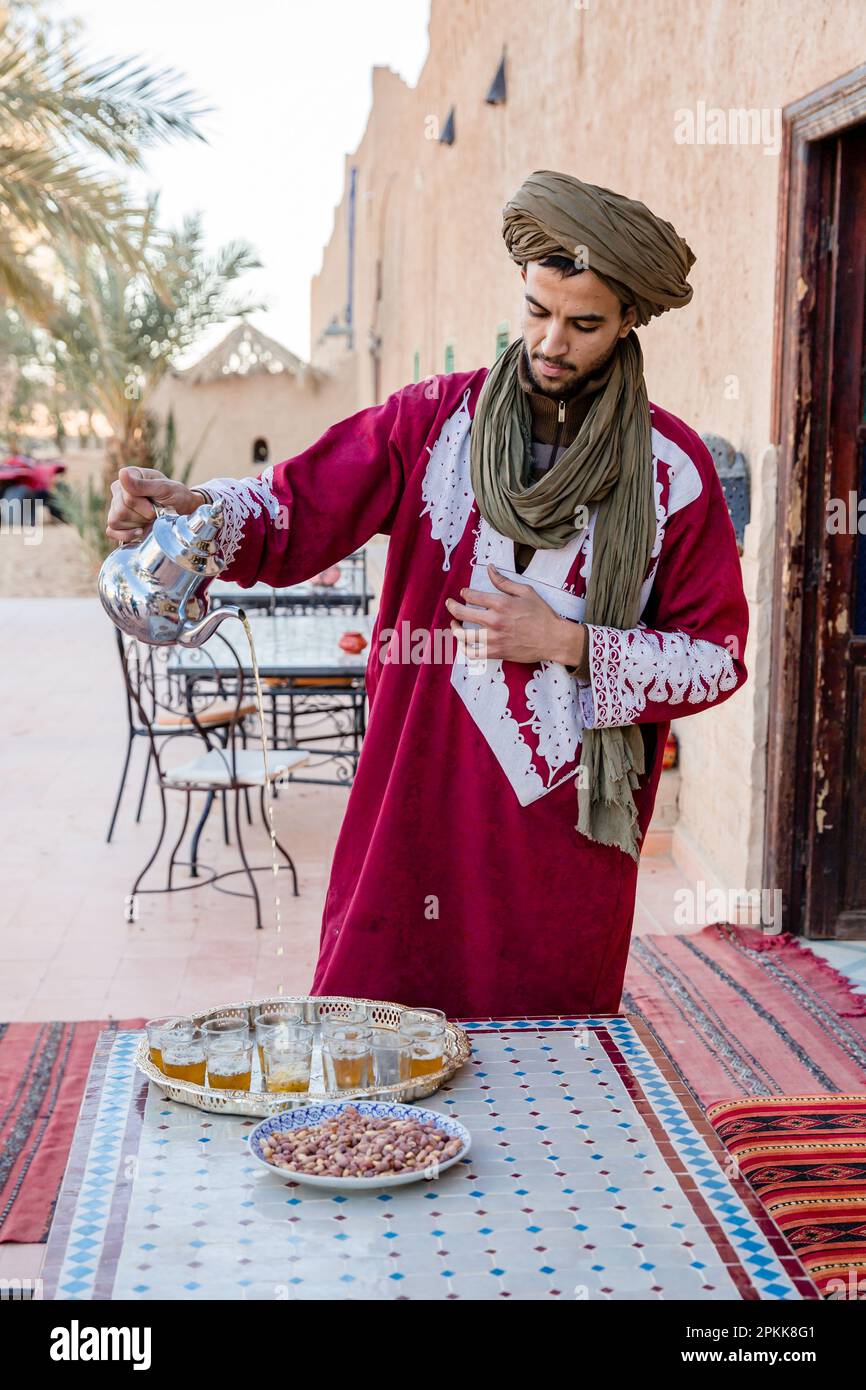 Ein traditionell gekleideter marokkanischer Mann schenkt Gläser Pfefferminztee zu Snacks ein Stockfoto