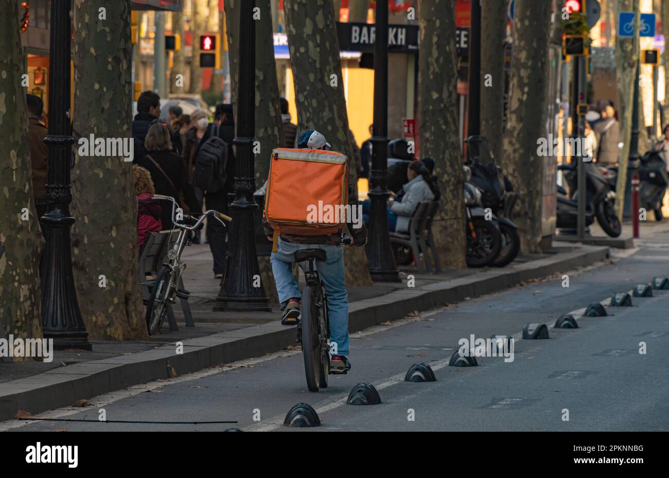 Ein Auslieferungsmann fährt mit einem orangefarbenen Rucksack auf einem Fahrradweg in Barcelona entlang einer von Bäumen gesäumten Straße, und Menschen gehen spazieren Stockfoto