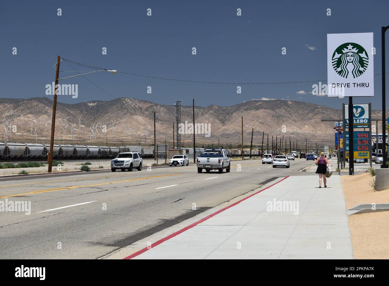 Blick vom Bürgersteig auf die Straße, Eisenbahnwaggons, Windturbinen und Berge: Blick nach Norden entlang Highway 14, Mojave, Kalifornien. Stockfoto
