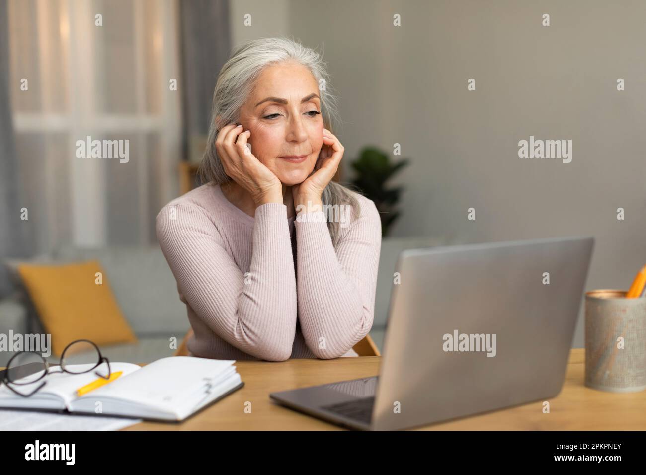 Fröhliche, hübsche, weiße Seniorin, die am Tisch sitzt, auf den Computer schaut, online telefoniert, sich Videos ansieht Stockfoto