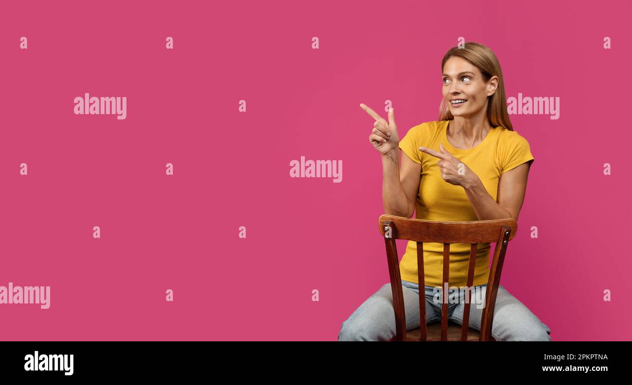 Fröhliche blonde Dame mittleren Alters, die auf einem Stuhl sitzt und mit dem Finger auf einen leeren Platz zeigt Stockfoto