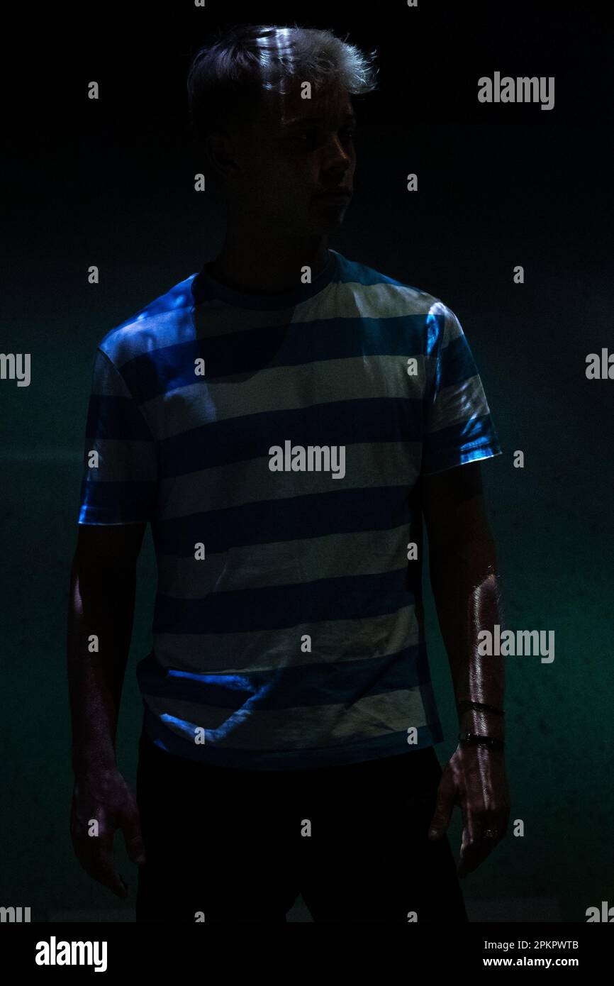 Ein Mann in einem gestreiften T-Shirt steht in einem dunklen Raum mit einem weichen weißen und blauen Licht, das auf ihn fällt Stockfoto
