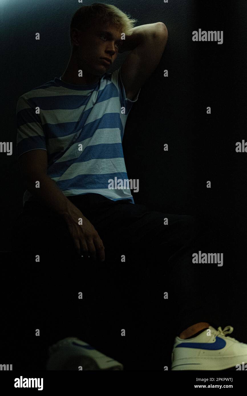Ein denkender Mann in einem gestreiften T-Shirt sitzt in einem dunklen Raum, in dem ein weiches natürliches Licht auf ihn fällt Stockfoto