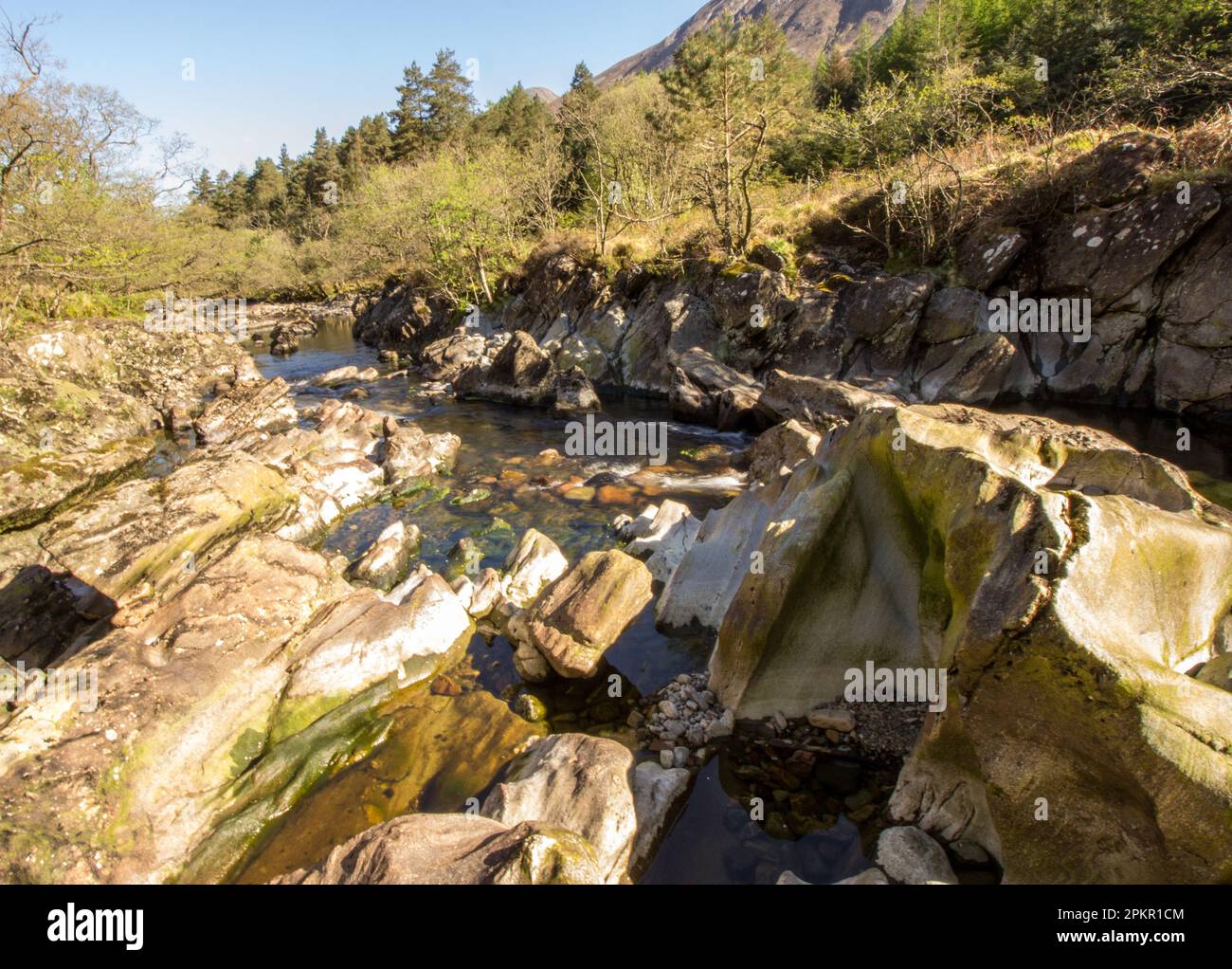 Der Fluss Coe, der an einem klaren, sonnigen Tag in den schottischen Highlands durch einen großen vulkanischen Felsen neben einem Torr fließt Stockfoto