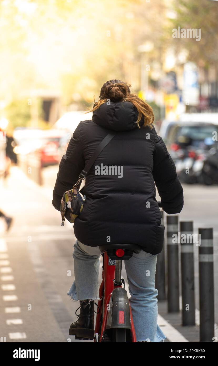 Junge Frau in einer schwarzen Jacke und Leggings auf dem Rücken, die auf einem roten Leihfahrrad auf einem Fahrradweg mit sonnendurchfluteten grünen Bäumen fährt Stockfoto