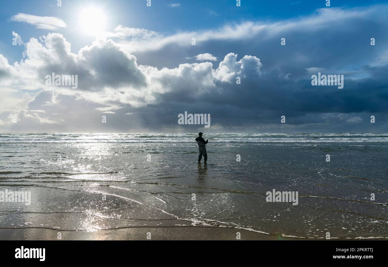 Ein alleinstehender Fischer in Neoprenanzug steht im Wasser der Ebbe und wird vom Sonnenlicht beleuchtet Stockfoto