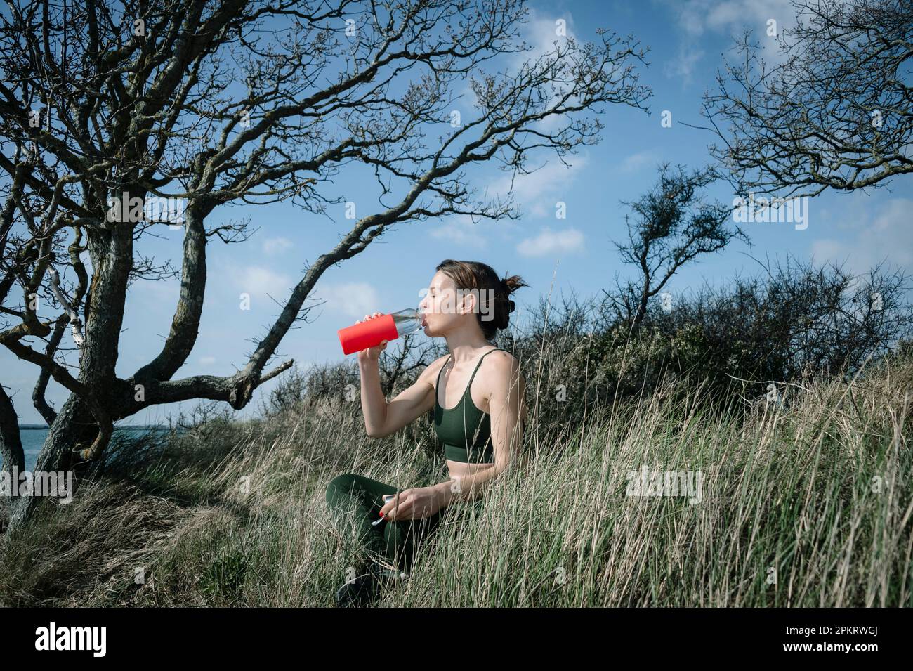 Eine sportliche Frau sitzt nach einem Training draußen auf dem Land und trinkt ein Glas Wasser. Stockfoto