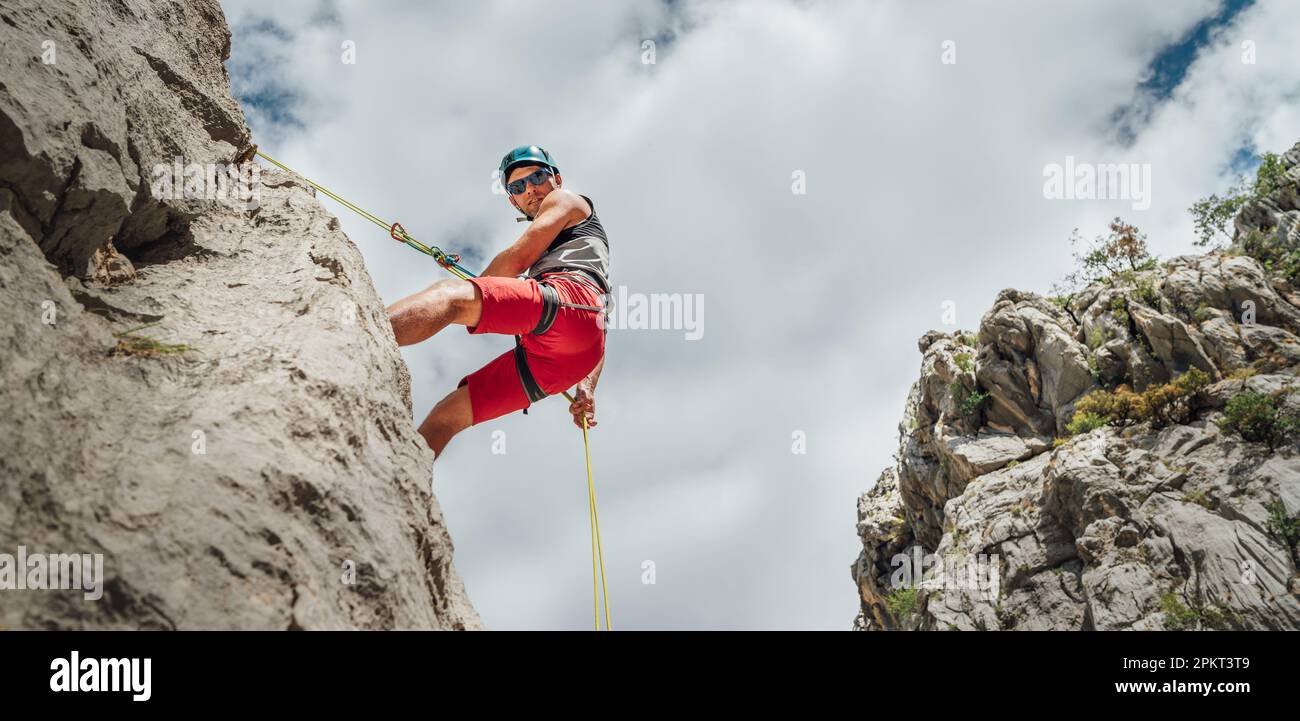 Aktiver Bergsteiger Mann mittleren Alters in Schutzhelm Blick auf Kamera, während Abseilen von Felswand mit Seil mit Sicherungsvorrichtung und Klettern ha Stockfoto
