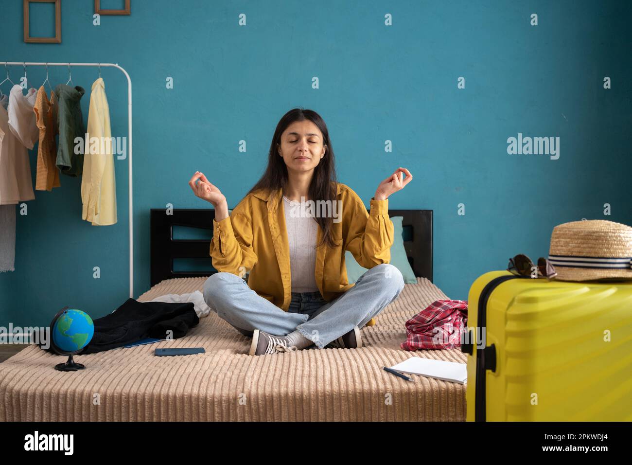 Junge Reisende Frau, die zu Hause neben dem Koffer auf dem Bett meditiert, Händchen in Yoga-Geste halten. Weiblicher Passagier, der am Wochenende ins Ausland reist Stockfoto