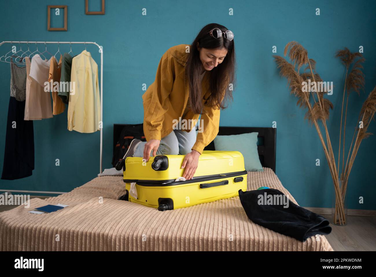 Vorbereitung des Reisekoffers zu Hause auf dem Bett. Eine junge Lateinin, die versucht, ihre Kleider in den gelben Koffer zu packen, um sich auf die Reise im Sommer vorzubereiten. Stockfoto