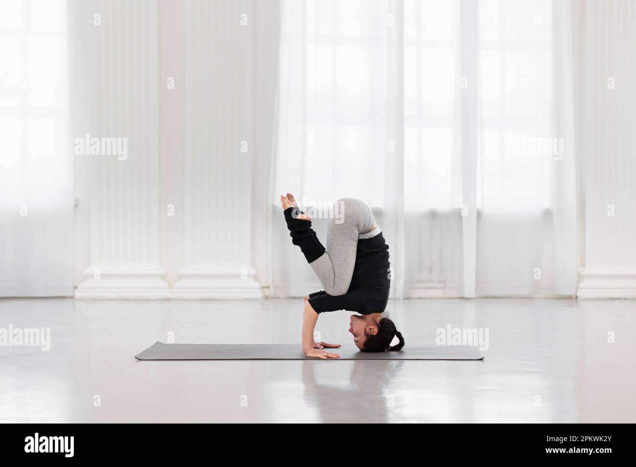 Seitenansicht einer Sportlerin, die während der Yoga-Meditation in einem hellen Fitnessstudio Kopfständertraining macht. Konzept der Normalisierung aller Prozesse im Körper Stockfoto