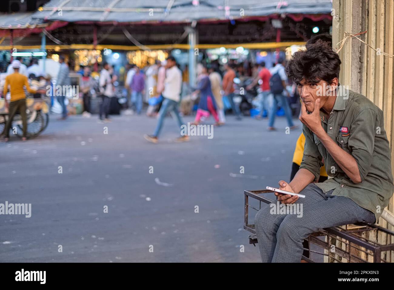 Ein junger Indianer sitzt aber penibel oder tief, reibt sich das Kinn und Handy in die Hand; in einer Straße in der Nähe von Crawford Market, Mumbai, Indien Stockfoto