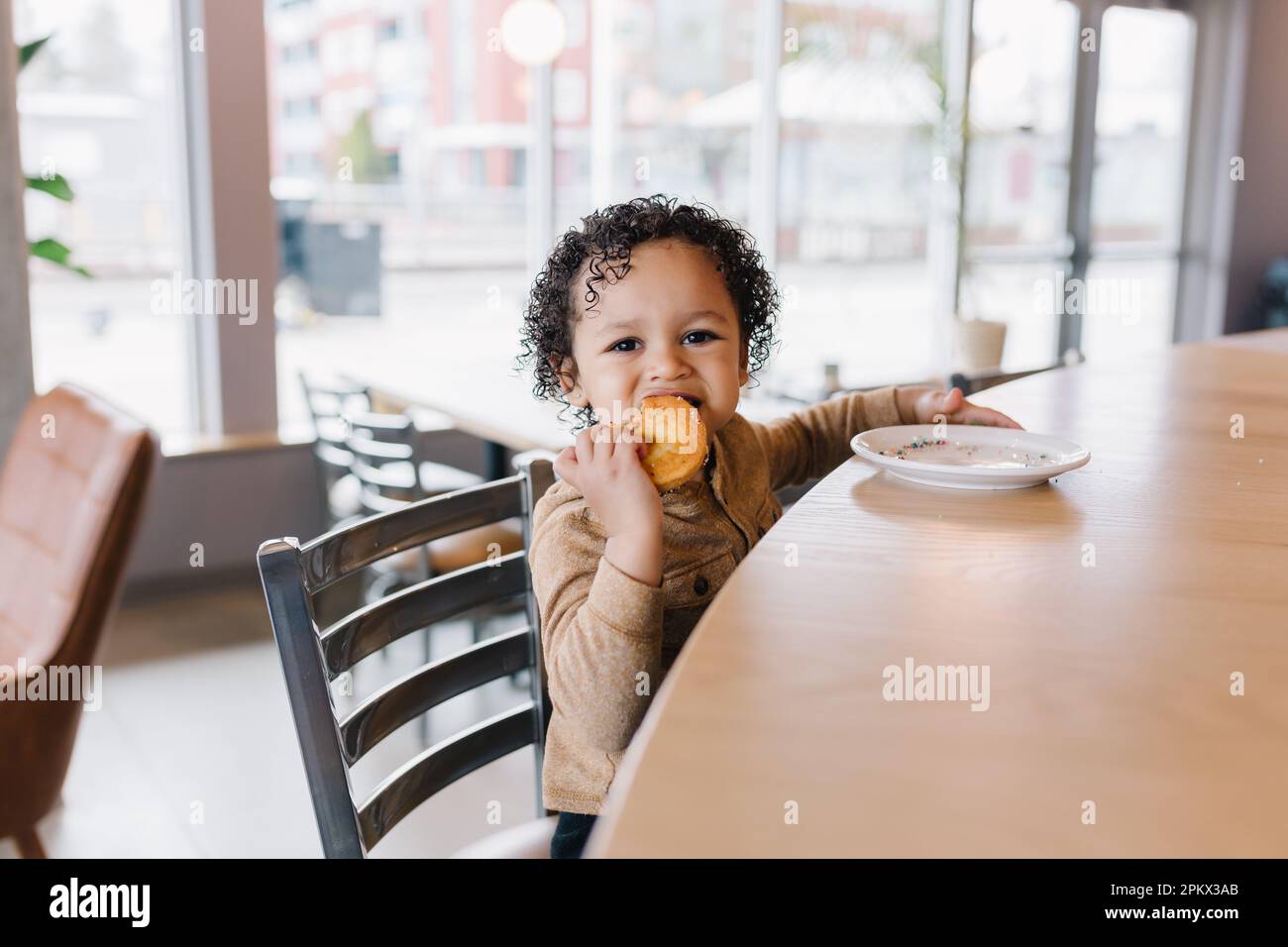 Ein bezauberndes, lockiges Kleinkind, das einen Donut im Café isst Stockfoto