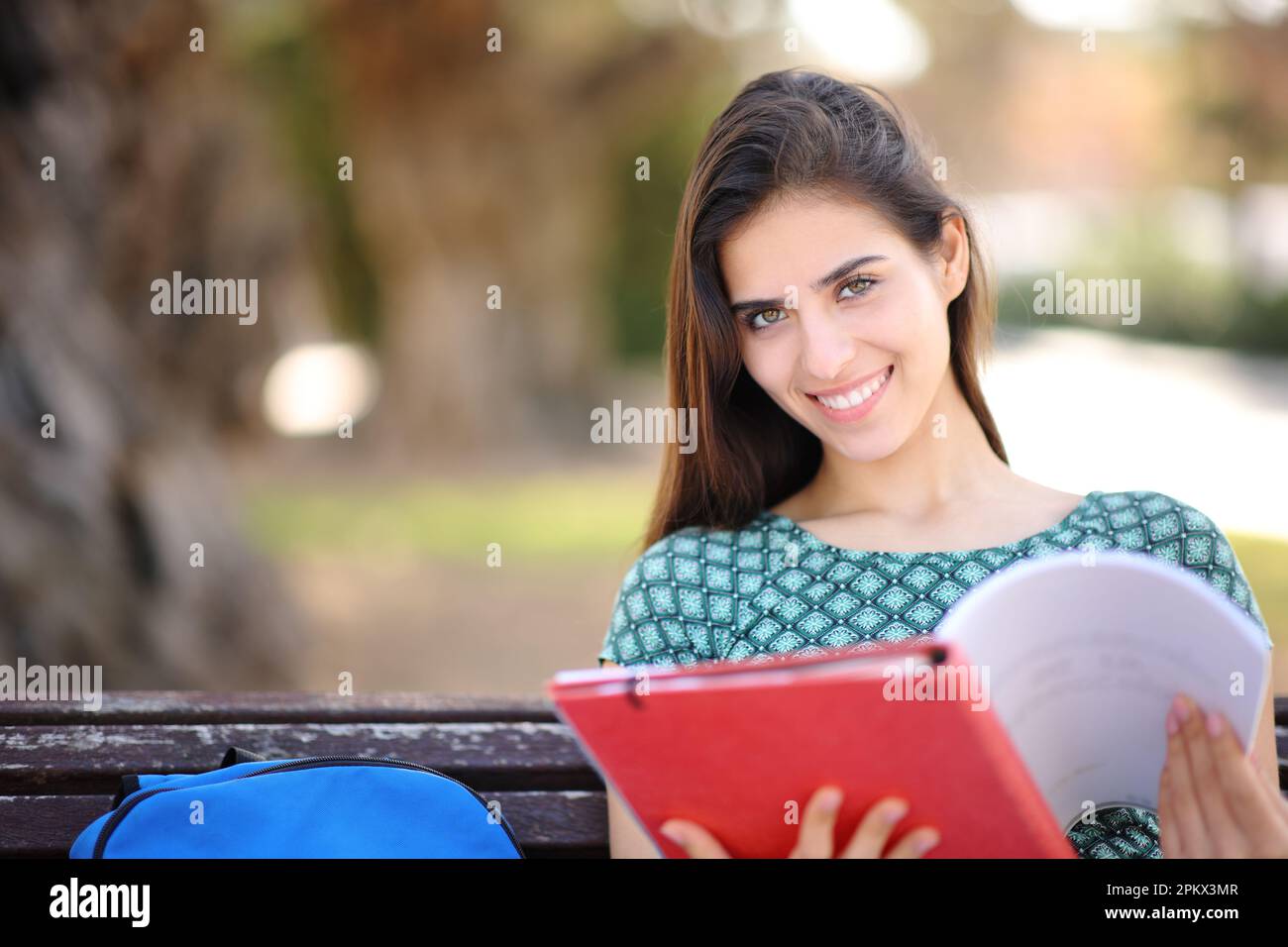 Glückliche Schülerin, die Notizen hält, schaut auf die Kamera, die auf einer Bank im Park sitzt Stockfoto