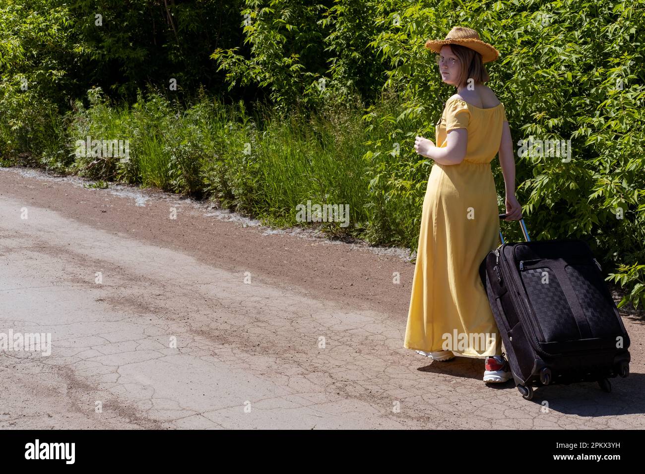 Asiatische Teenagerin in gelbem Kleid, Brille und Hut, die mit einem Koffer auf einer Landstraße spaziert. Ökotourismus. Sommerzeit. Feiertage. Anfahrhilfe Stockfoto