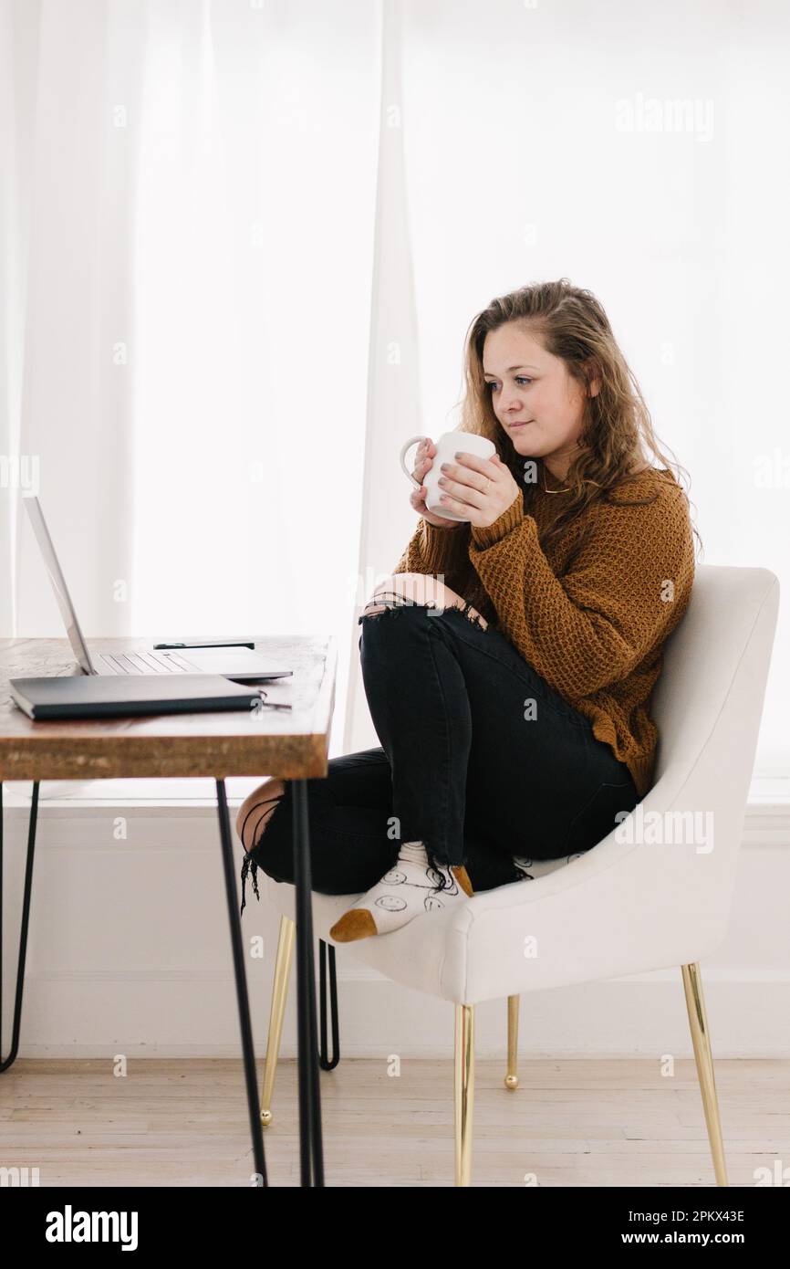 Junge Frau, die auf einen Computerbildschirm schaut, sich auf dem Stuhl aufgerollt hat, einen Becher in der Hand hält Stockfoto