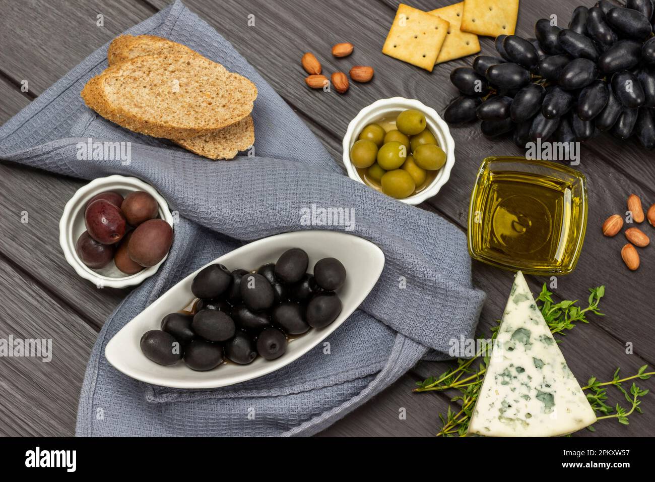 Schwarze Oliven in einer Schüssel und ein Haufen schwarze Trauben. Blauschimmelkäse und Nüsse. Graue Serviette. Flach verlegt. Dunkler Holzhintergrund. Stockfoto
