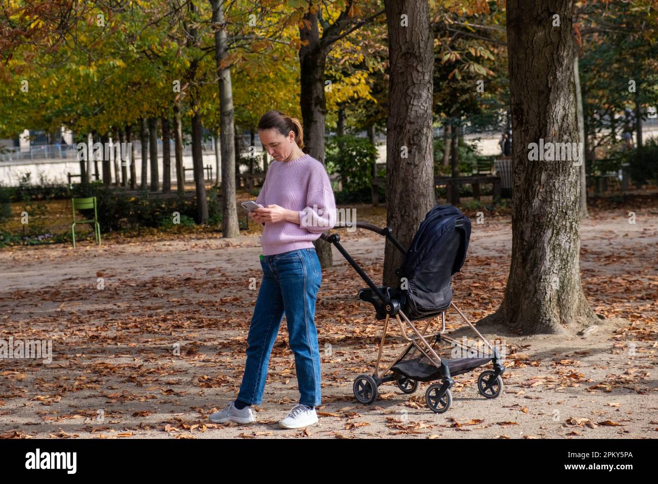 Mutter, die ihr Handy mit dem Kinderwagen im malerischen Pariser Garten überprüft Stockfoto