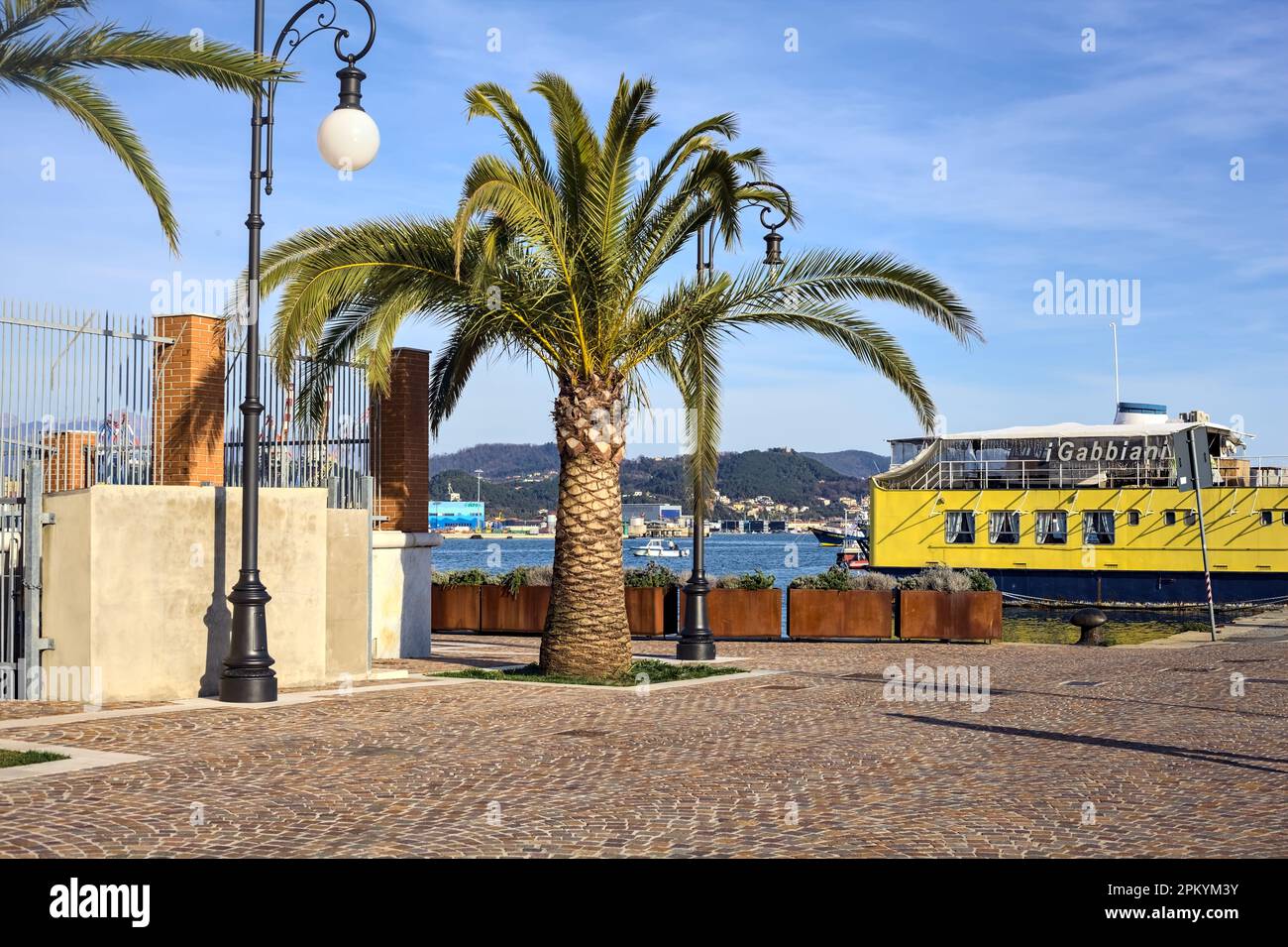 Palme und eine Straßenbeleuchtung auf einem Pier mit einem gelben Schiff dahinter bei Sonnenuntergang Stockfoto