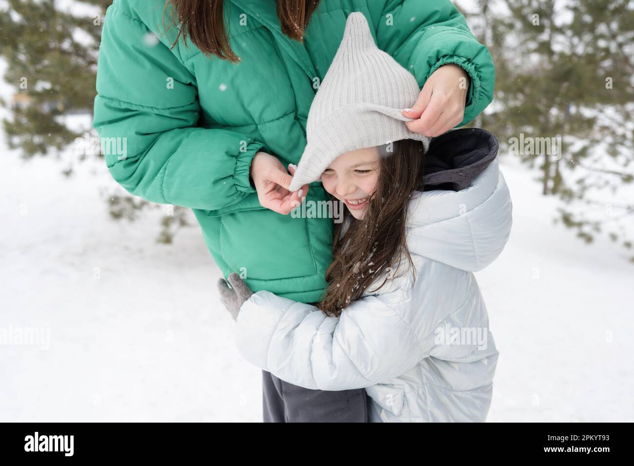 Gekürzte, nicht wiedererkennbare Frau in warmer Kleidung, einstellende Mütze der umarmenden jungen Tochter und im Winterpark vor verschwommenen Bäumen stehen Stockfoto