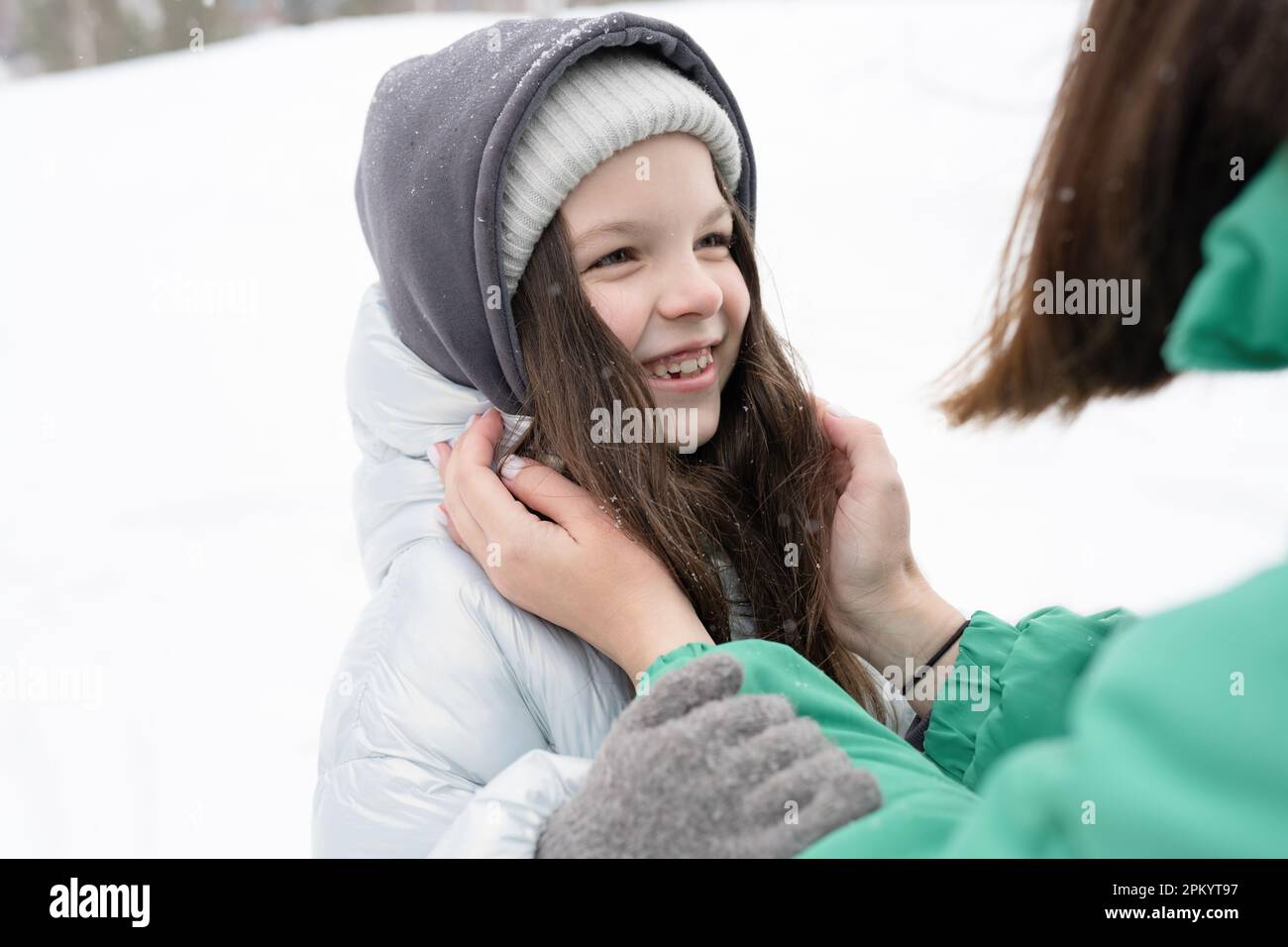 Gekürzte, nicht wiedererkennbare Frau in warmer Kleidung, einstellende Mütze der umarmenden jungen Tochter und im Winterpark vor verschwommenen Bäumen stehen Stockfoto