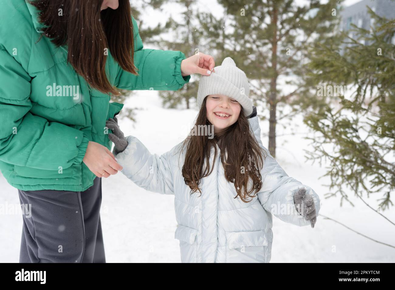 Gekürzte, nicht wiedererkennbare Frau in warmer Kleidung, einstellende Mütze der umarmenden jungen Tochter und im Winterpark vor verschwommenen Bäumen stehen Stockfoto
