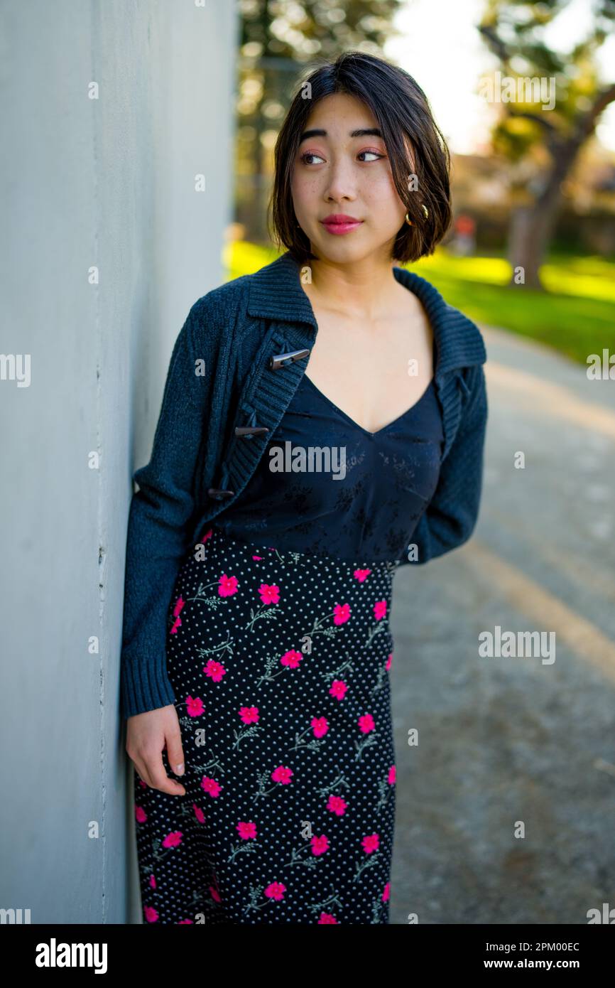 Junge asiatische Frau lehnt sich zur Goldstunde gegen eine graue Betonwand | warme Farbtöne | Rock mit Blumenmuster | Oberteil aus schwarzer Seide Stockfoto