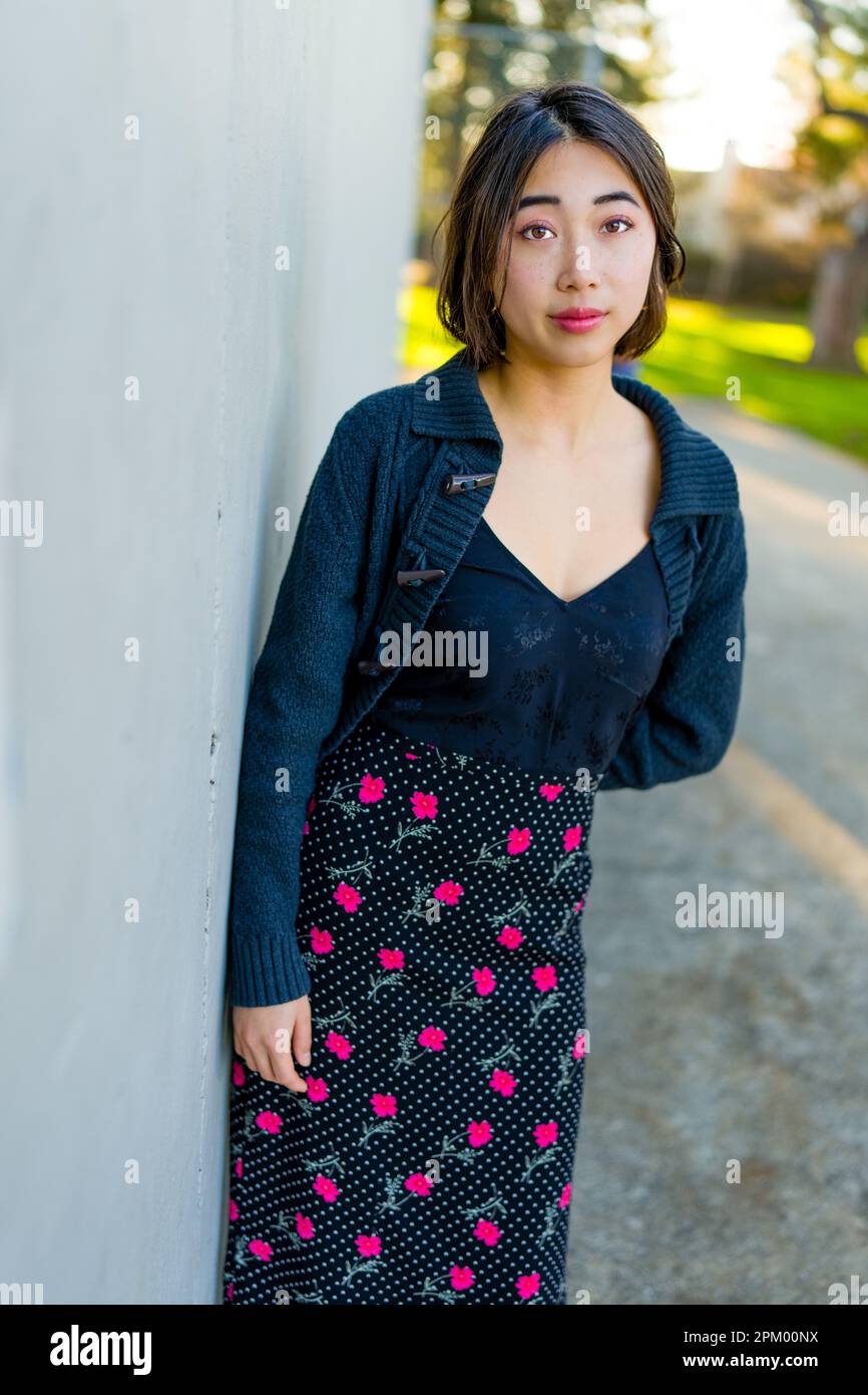 Junge asiatische Frau lehnt sich zur Goldstunde gegen eine graue Betonwand | warme Farbtöne | Rock mit Blumenmuster | Oberteil aus schwarzer Seide Stockfoto