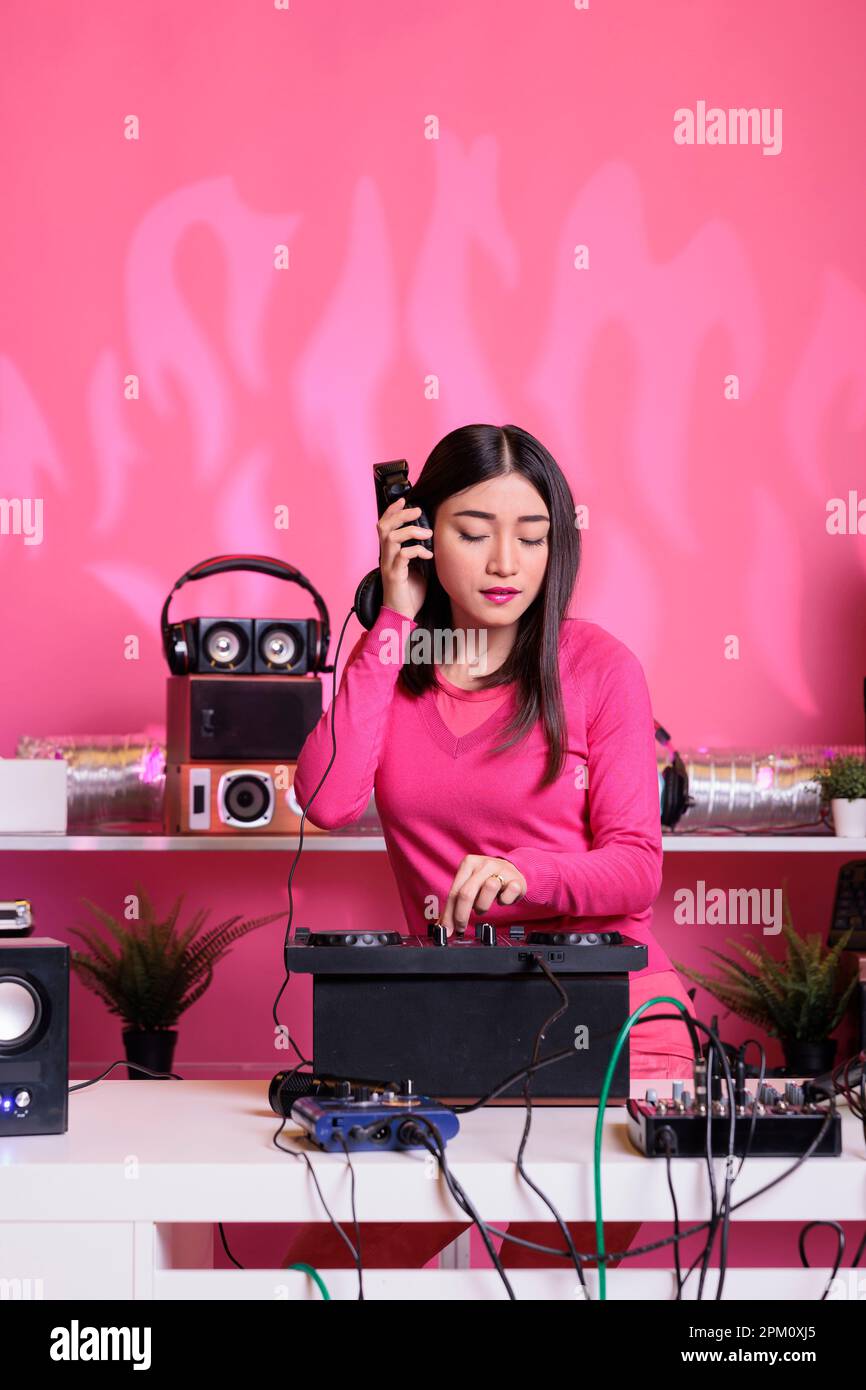 Asiatischer Künstler mit Kopfhörern, der Spaß hat, während er Techno-Musik  über eine professionelle Mixerkonsole spielt, steht am dj-Tisch im Studio  mit pinkfarbenem Hintergrund. Musiker, der im Club ein elektronisches Lied  spielt Stockfotografie -