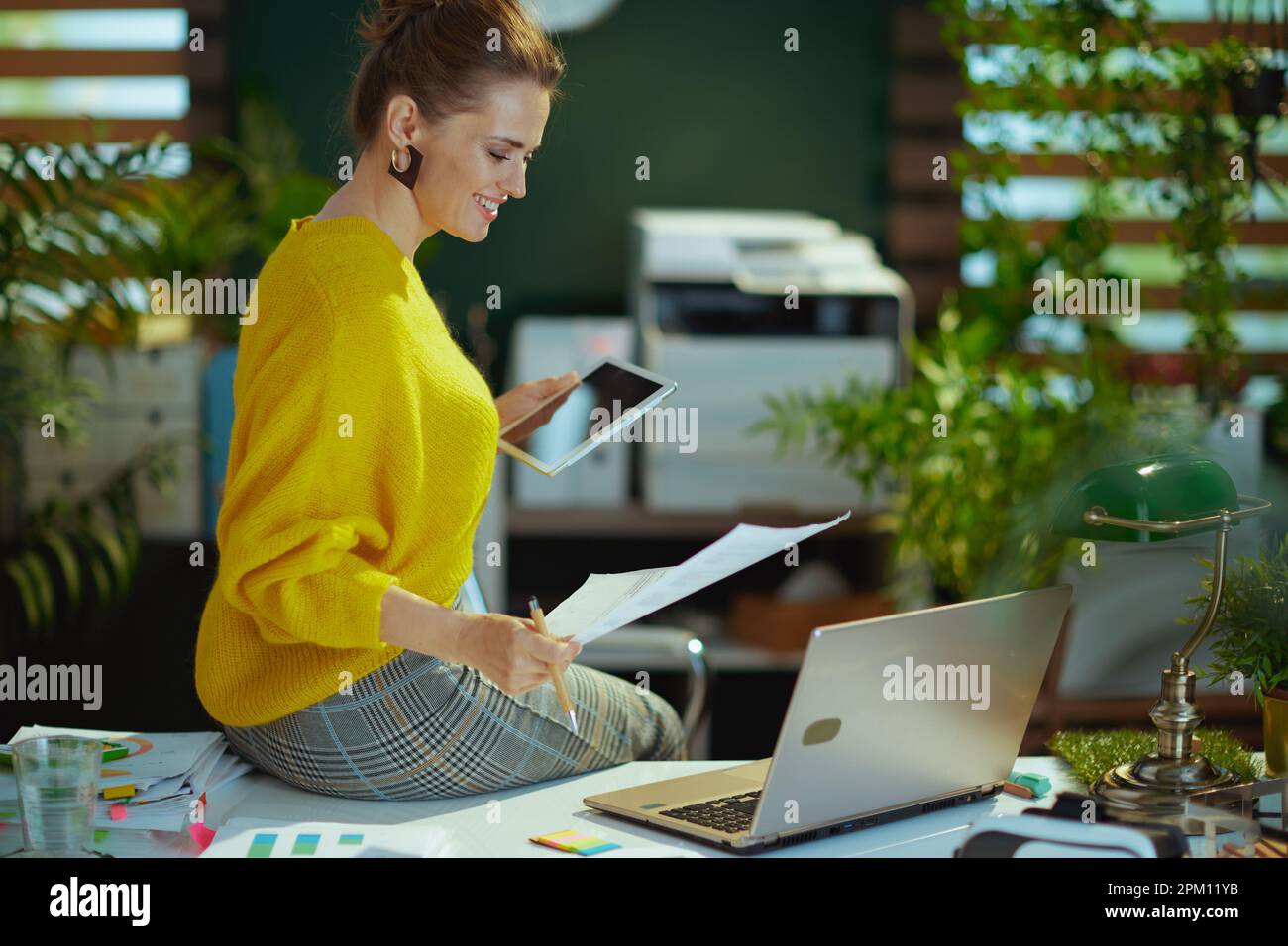 Glückliche, moderne Eigentümerin eines kleinen Unternehmens in einem gelben Pullover mit Tablet-PC und Laptop, die in einem modernen grünen Büro mit Dokumenten arbeiten. Stockfoto