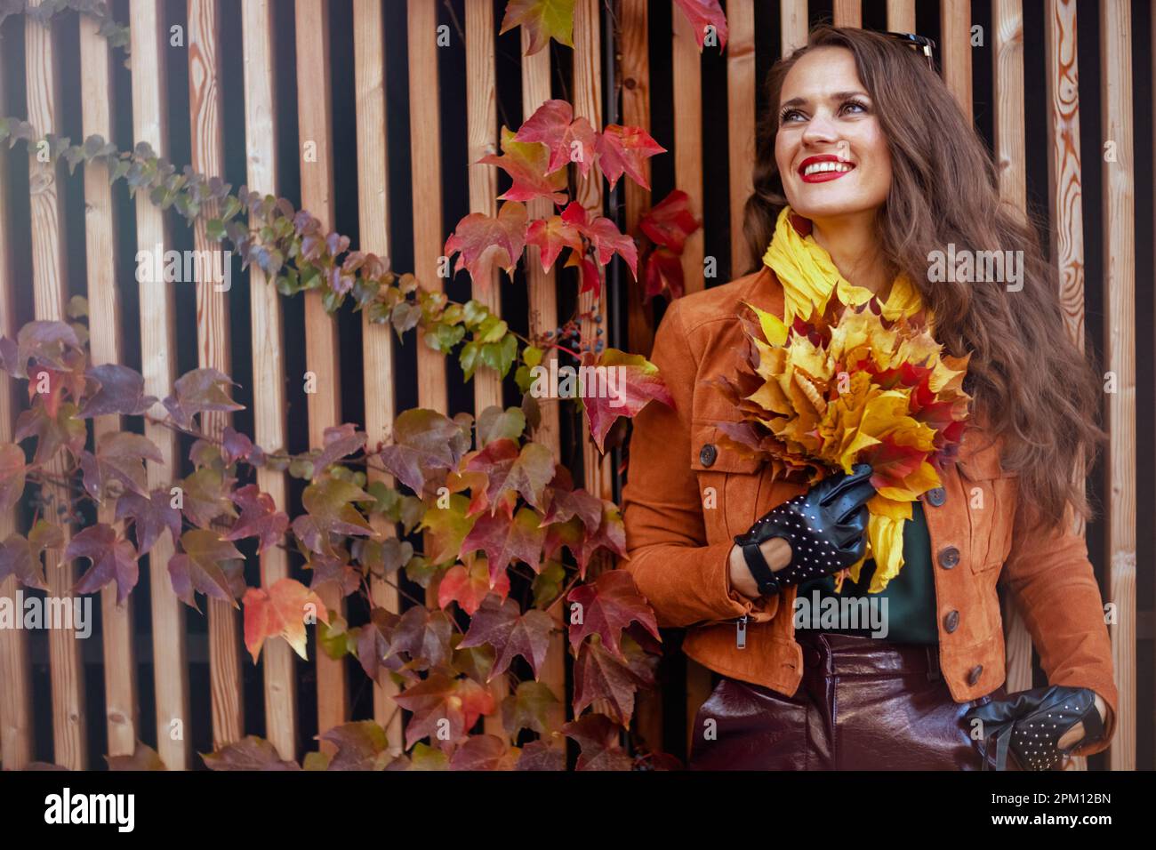 Hallo november. Entspannte, trendige 40-jährige Frau in orangefarbenem Trenchcoat mit herbstgelben Blättern in der Stadt. Stockfoto