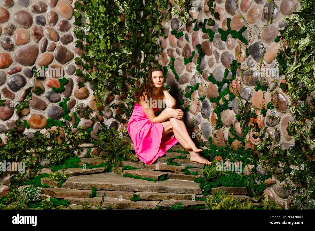 Eine Erwachsene Frau sitzt in ihrem süßen Sommerkleid mit welligen Haaren und einem bezaubernden Lächeln auf einem Steinteller in der Nähe der Wand. Sie sieht entspannt und zufrieden aus. Viel Spaß Stockfoto
