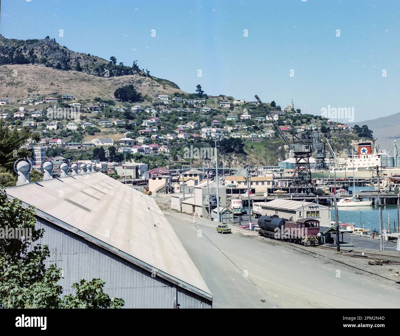 Ein historisches Bild aus dem Jahr 1981 von einem Güterzug, einer Eisenbahnlinie und Lagerhallen am Hafen von Lyttelton in der Nähe der Stadt Christchurch auf der Südinsel Neuseelands. An der Anlegestelle liegt eines der Blue Star Line, Frachtschiff und im Vordergrund befinden sich stark Bros. Trucks. Stockfoto