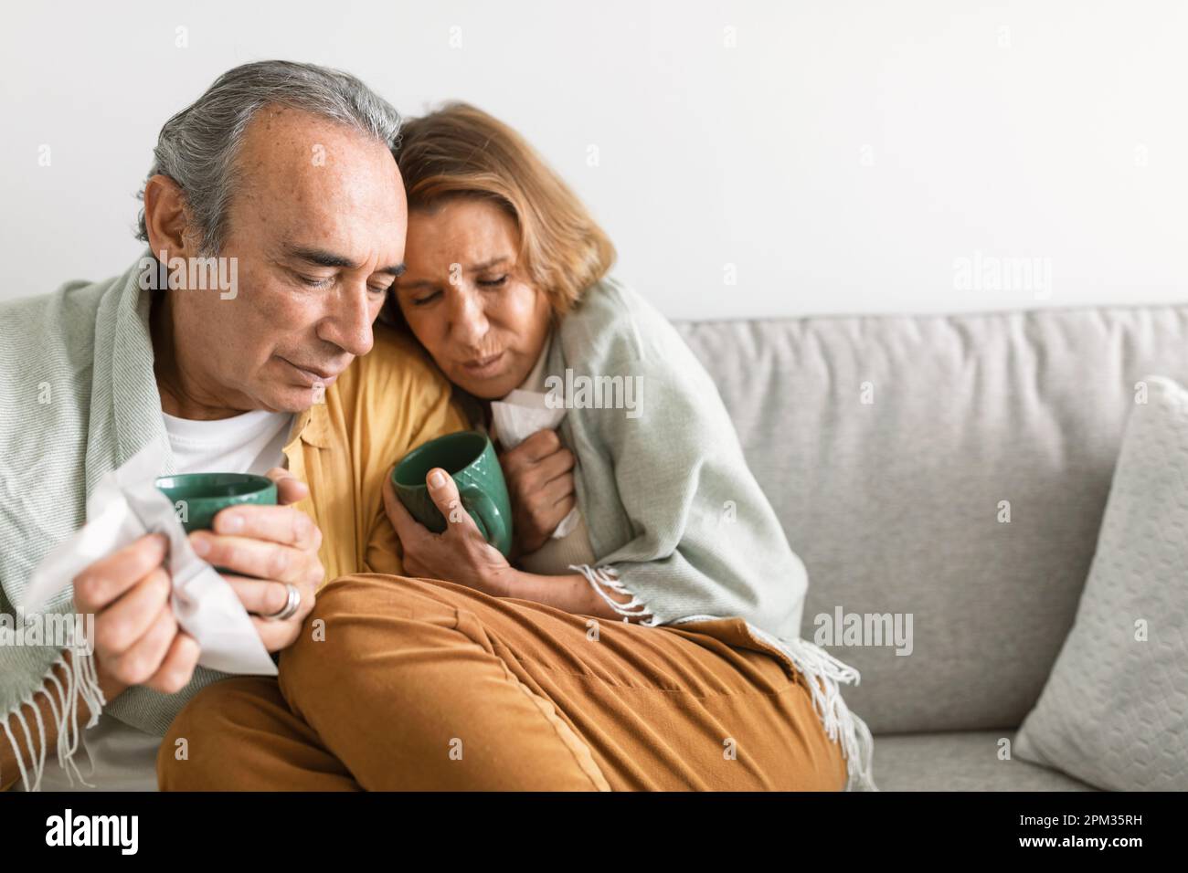 Ältere Ehepartner, die in eine Decke eingewickelt sind und Tassen mit heißem Tee oder Kaffee halten, sich krank und kalt fühlen, auf dem Sofa sitzen, freier Platz Stockfoto