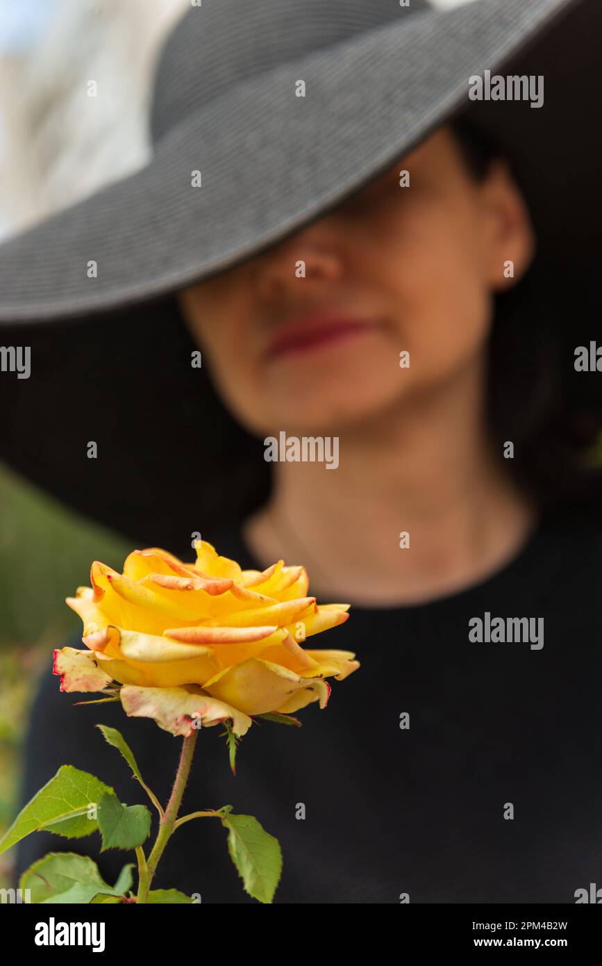 Verschwommenes Bild einer Frau mit Hut, die eine gelbe Rose bewundert, selektiver Fokus. Stockfoto