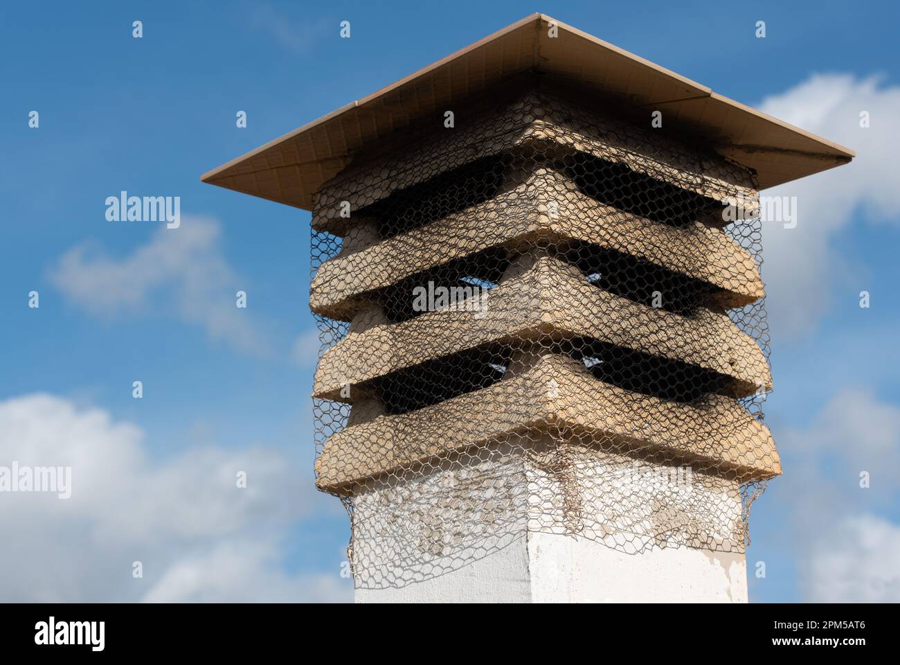 Nahaufnahme eines alten Schornsteins vor einem blauen Himmel. Da ist ein Dach auf dem Kamin. Seitlich ist ein Drahtgeflecht angebracht, um ein Einfliegen von Vögeln zu verhindern. Stockfoto