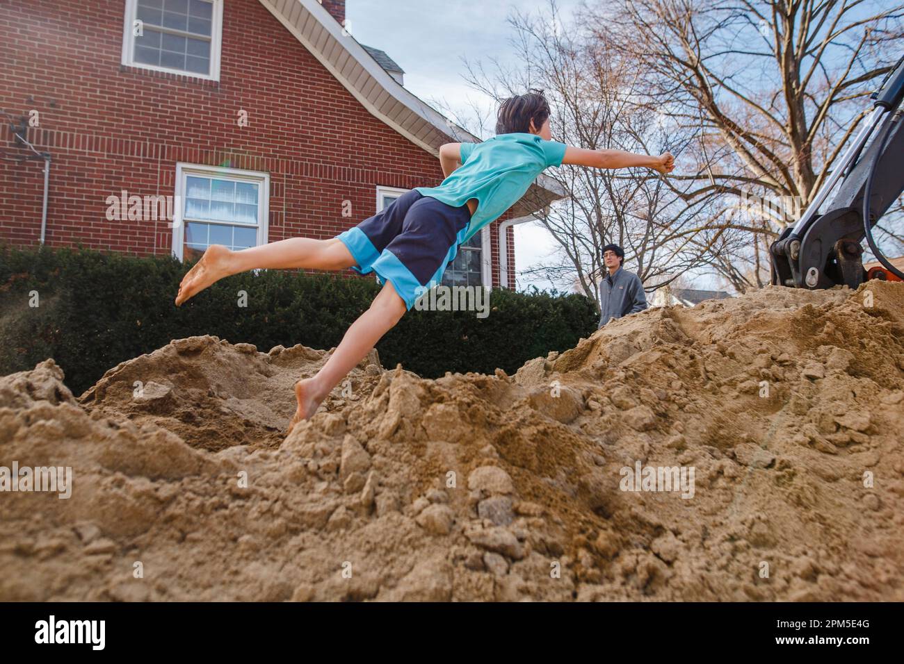 Ein Barfußjunge springt in einen riesigen Sandhaufen, während der Vater zusieht Stockfoto