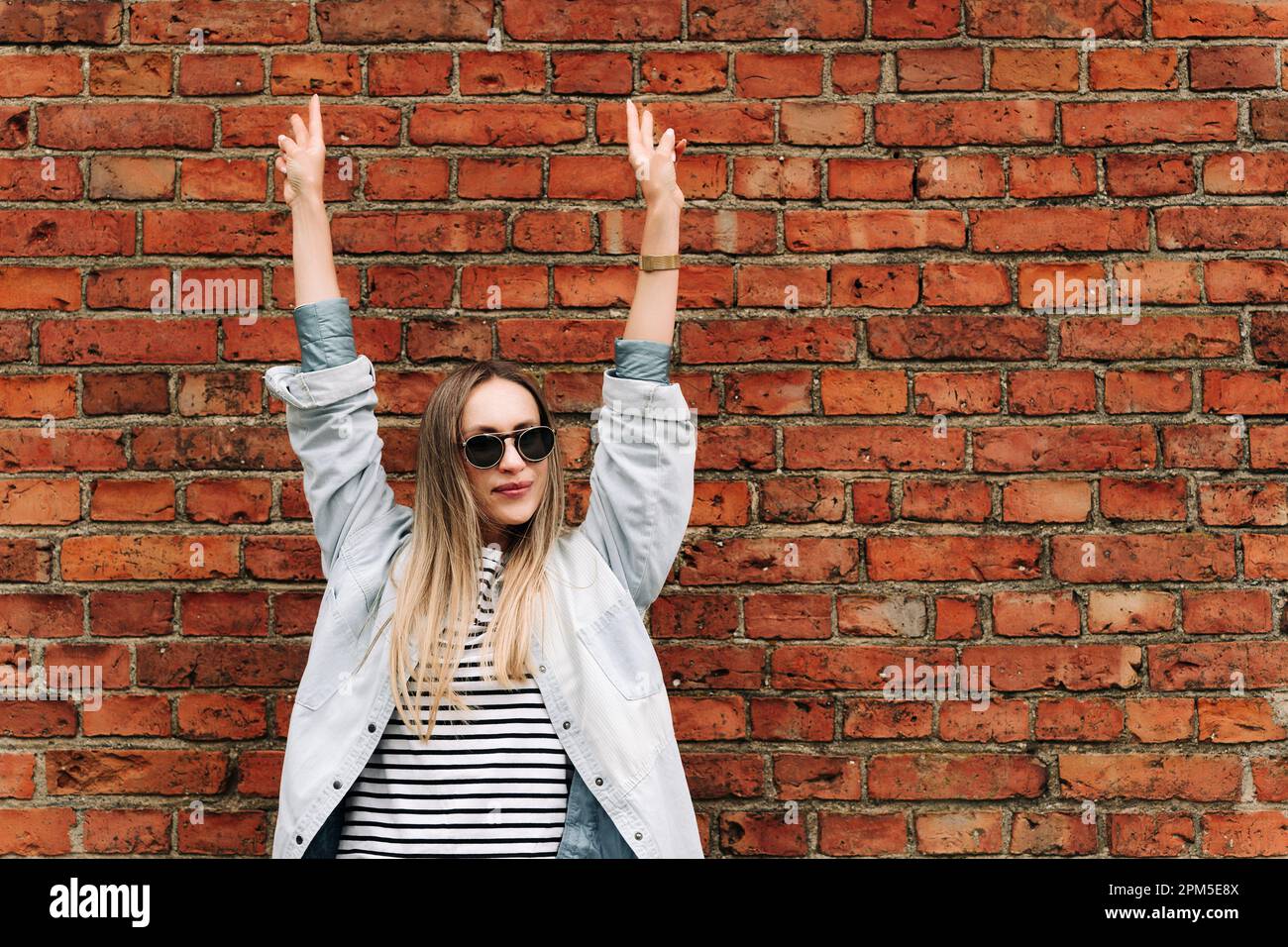 Eine Frau mit Sonnenbrille, mit erhobenen Händen gegen eine Ziegelwand Stockfoto