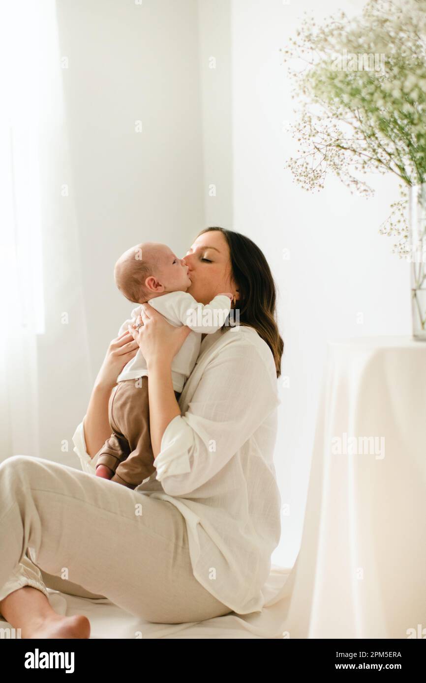 Sitzende junge Mutter küsst den neugeborenen Sohn am Fenster und Blumen Stockfoto
