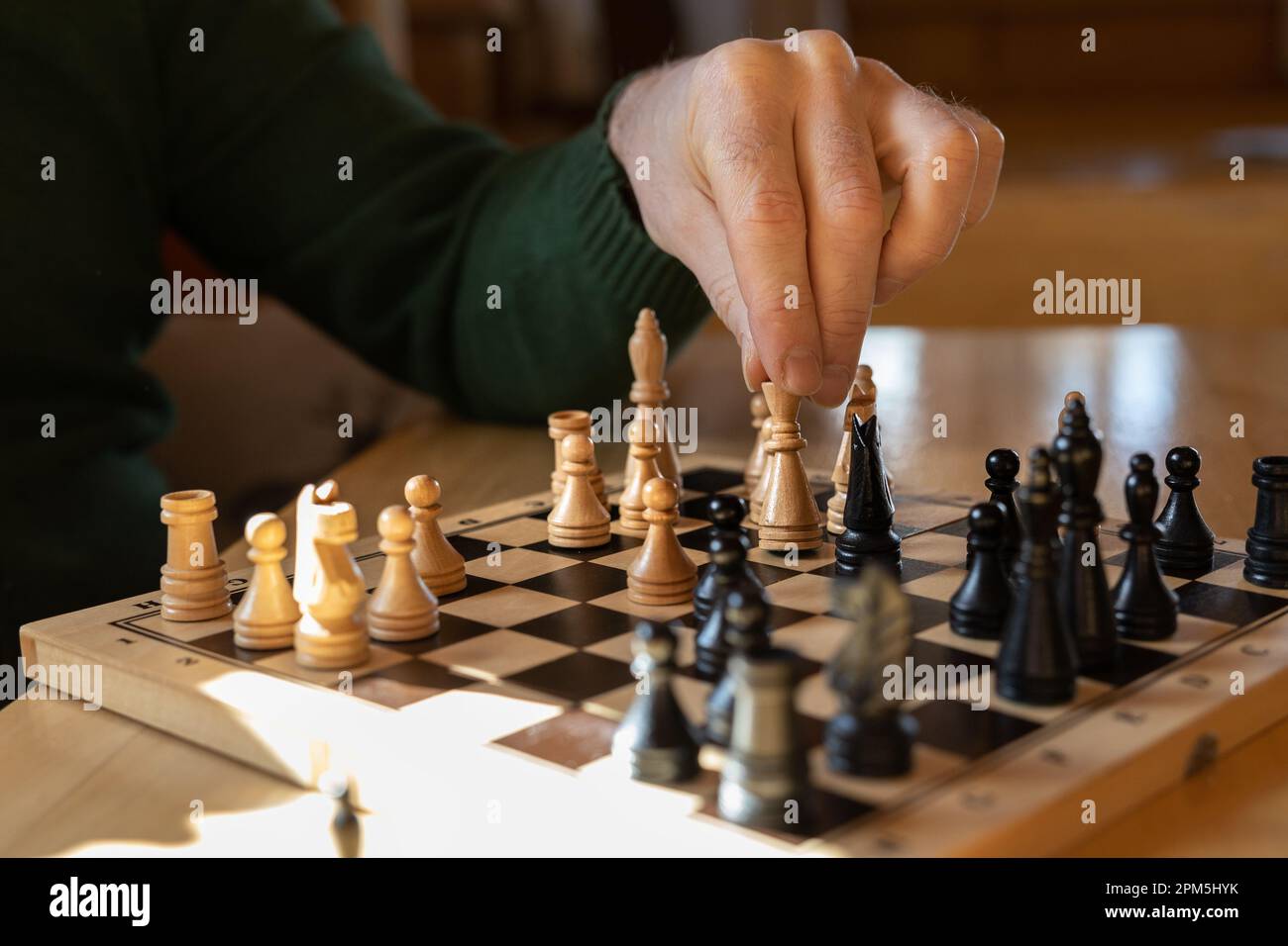 Ein Mann sitzt an einem Holztisch und macht eine Schachbewegung mit einem weißen Stück. Stockfoto