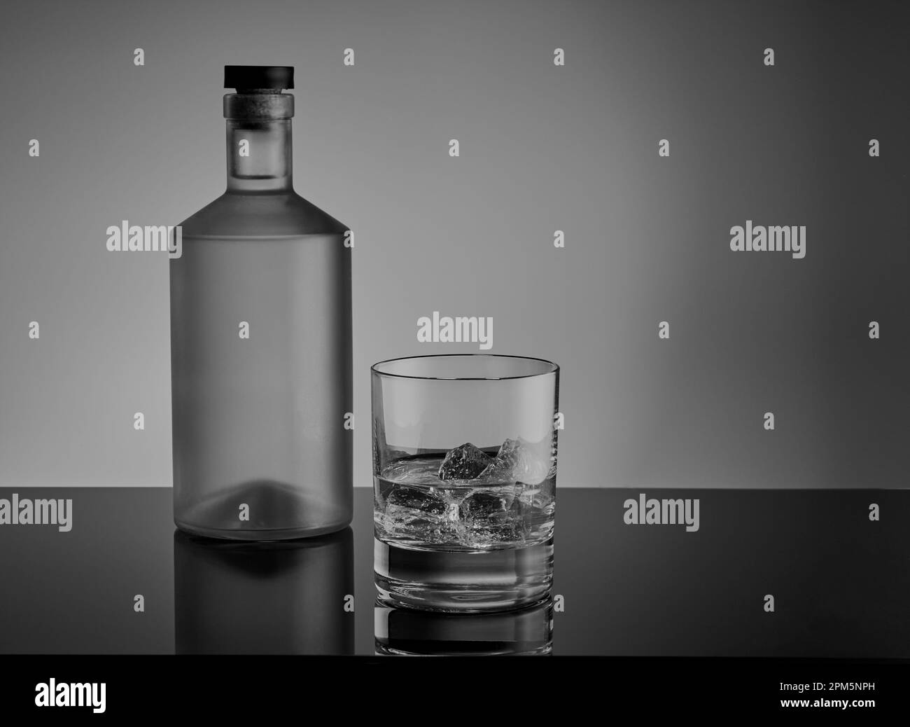 Schwarz-weiß-Bild einer Flasche Spirituosen mit einem Glas Whisky Tumbler mit Eis und Alkohol auf einer reflektierenden Oberfläche. Stockfoto