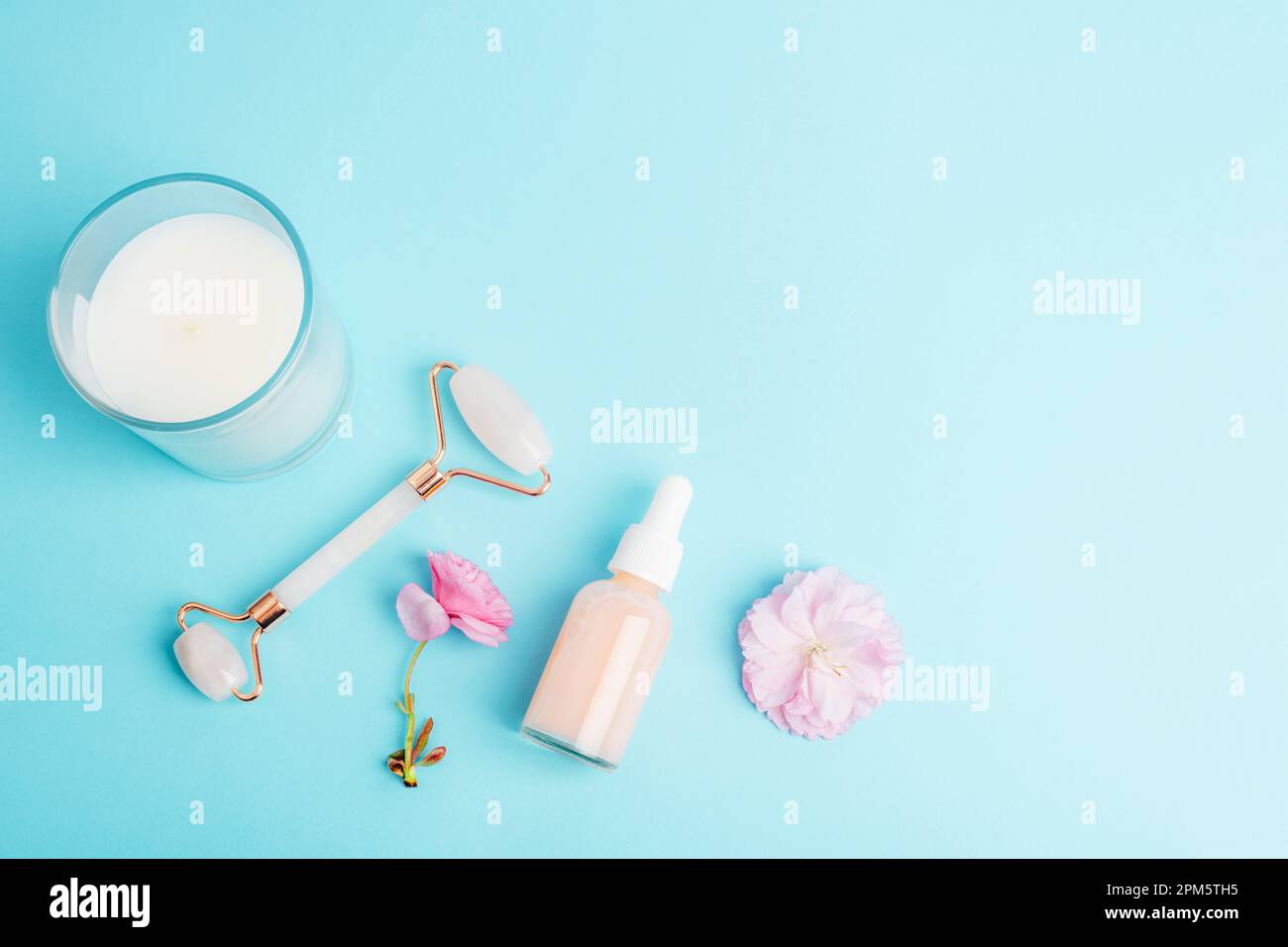 Serumflasche, Gesichtsrolle, Gua-Sha-Stein, Aroma-Kerze und rosafarbene Sakura-Blüte auf blauem Hintergrund. Hautpflege, Schönheitsbehandlungskonzept. Draufsicht, flach Stockfoto