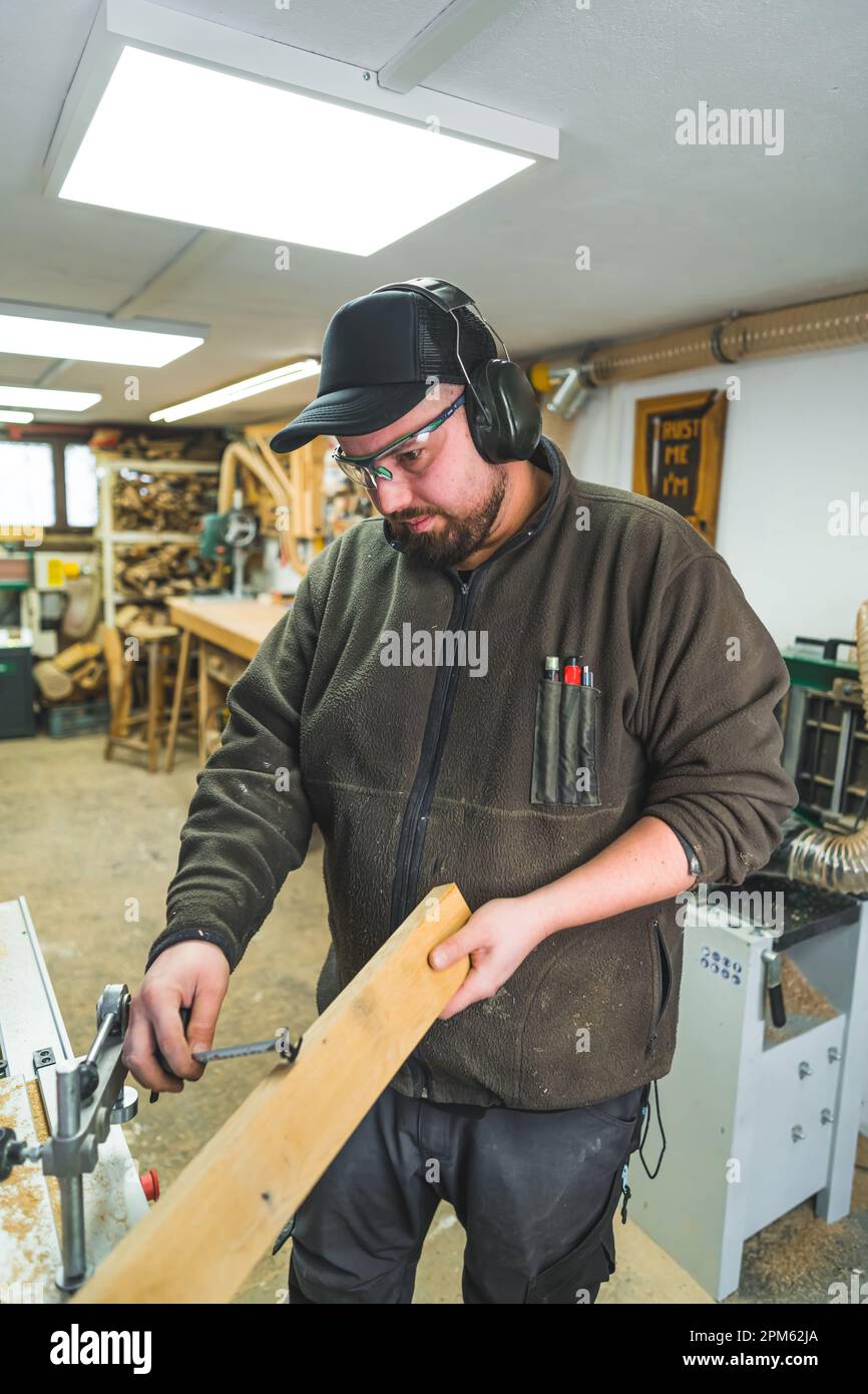 Professioneller Zimmermann in Kopfhörern und Brillen und hält eine Planke. Vertikaler Indoor-Shot. Holzverarbeitende Innenausstattung. Hochwertiges Foto Stockfoto