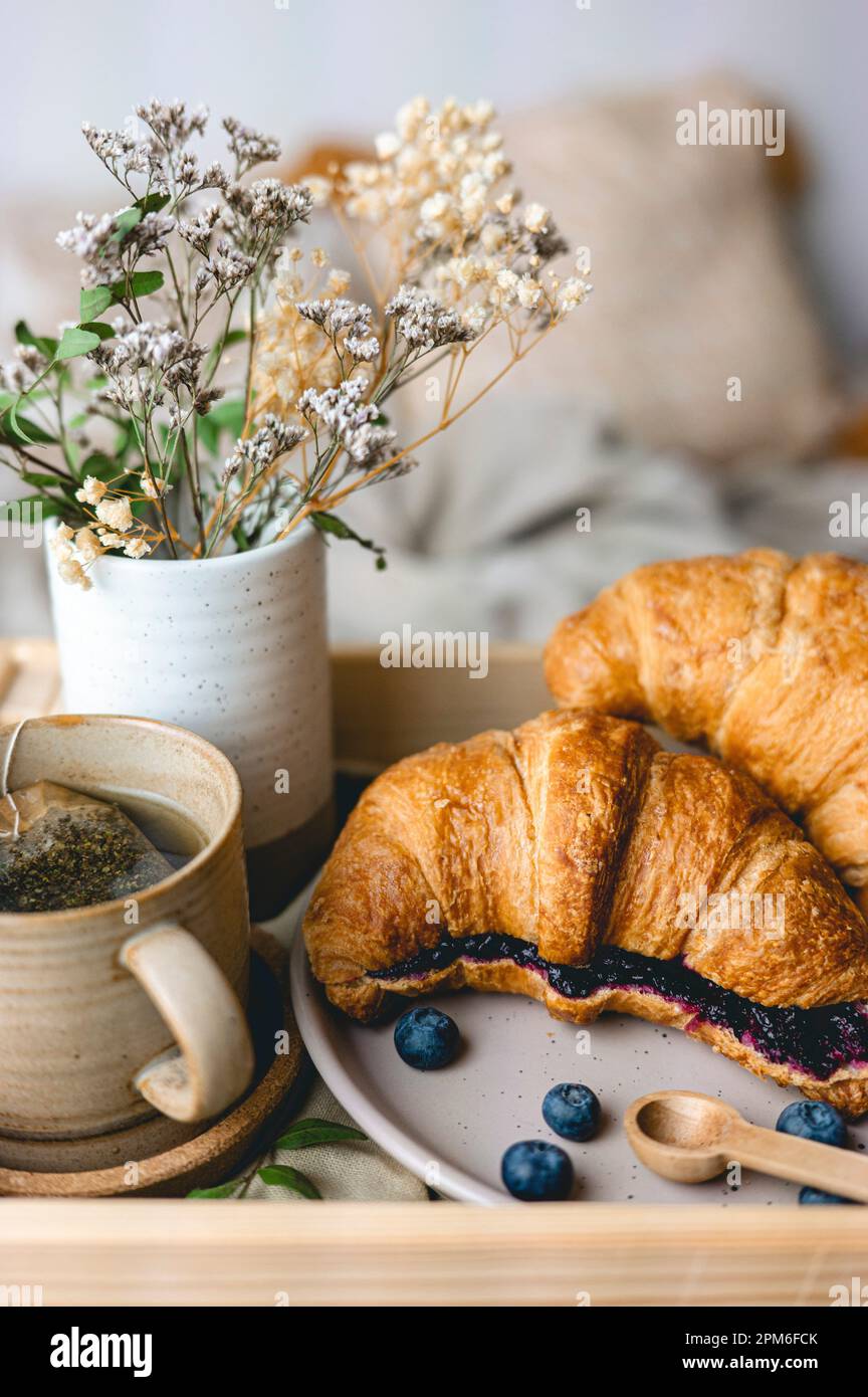 Frühstück im Bett mit frisch gebackenen Croissants mit Blaubeermarmelade und Tee in einer Tasse. Stockfoto