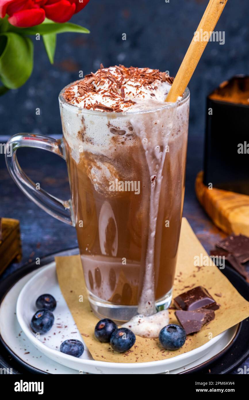 Mandelmilch heiße Schokolade mit Schlagsahne in einem Latte-Glas, Blaubeeren und Schokoladenstücke, Blumen dahinter. Stockfoto